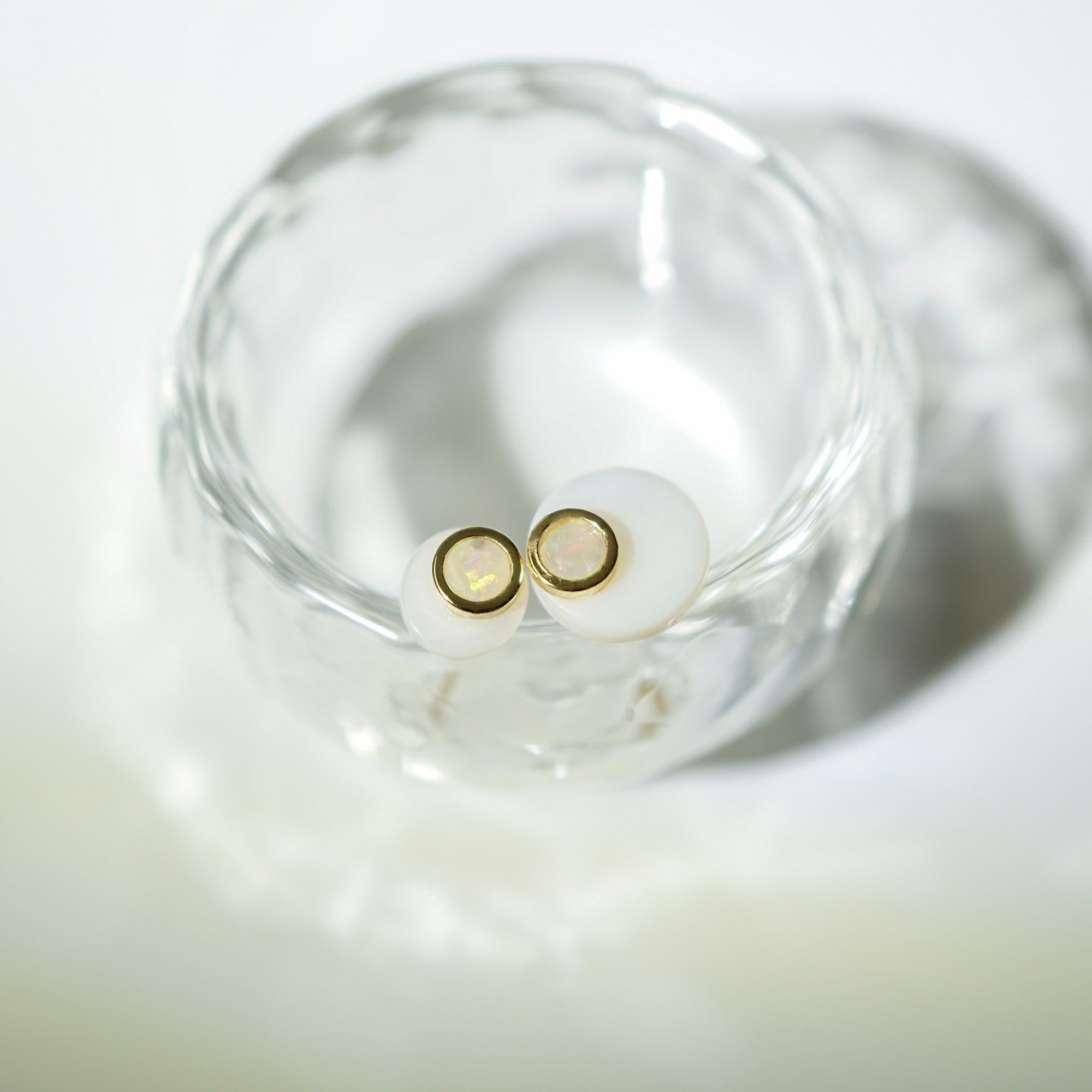A pair of small stud earrings with round white stones and thin gold settings, resting inside a clear glass dish on a soft white surface.