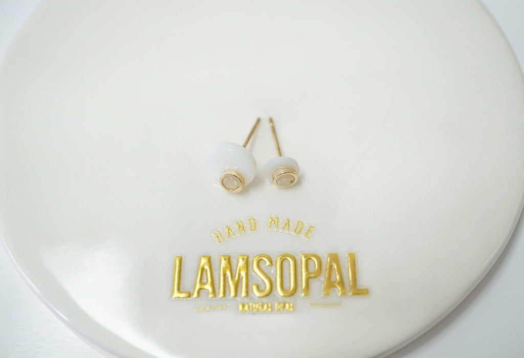 A pair of small white dome stud opal earrings with gold posts and bezel settings, displayed on a glossy white circular plate with gold 'LAMSOPAL HAND MADE' branding.