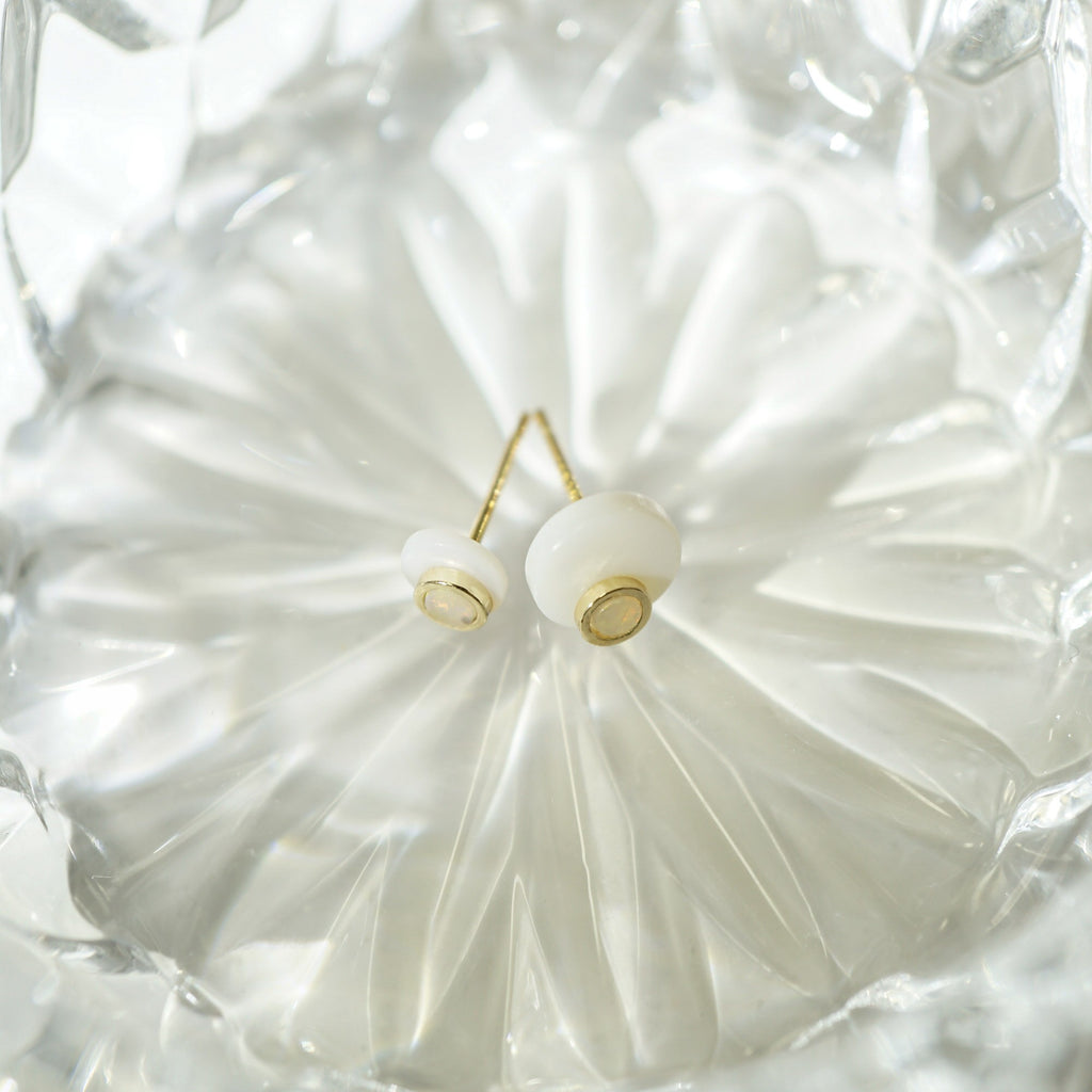 A pair of gold stud opal earrings with round white beads, displayed on a textured, faceted clear glass dish.