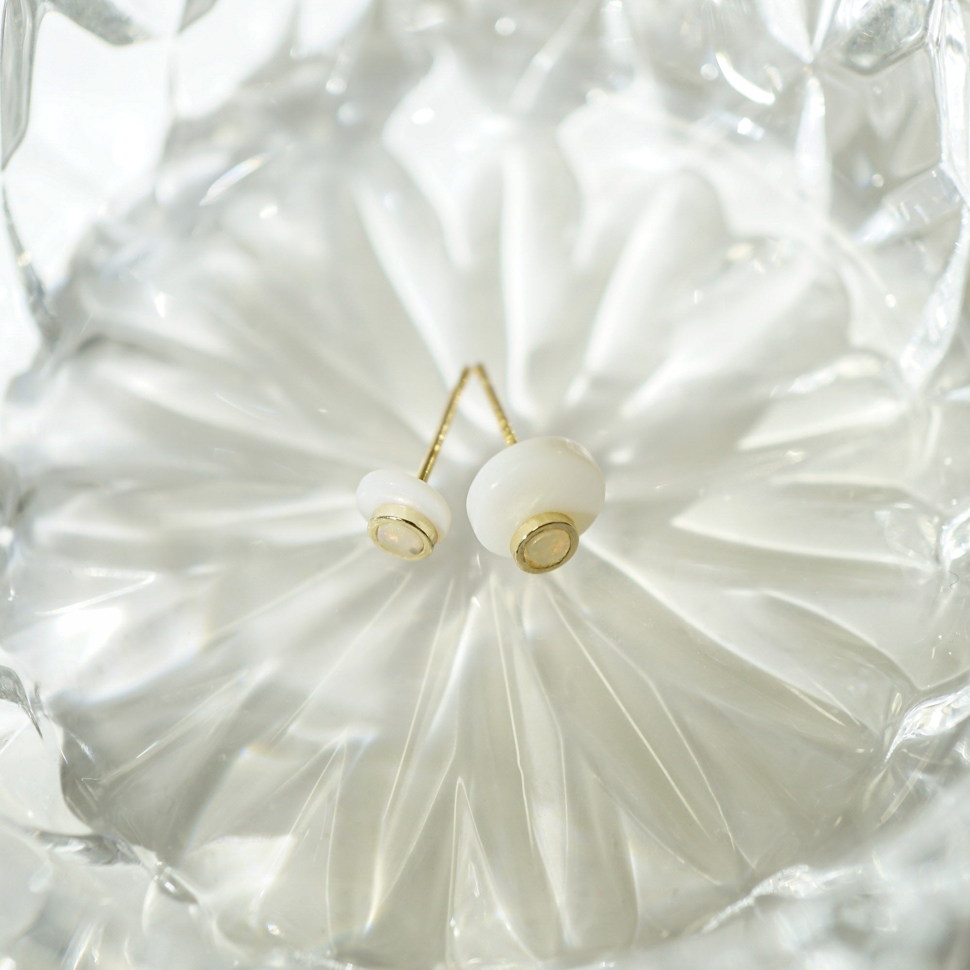 A pair of gold stud opal earrings with round white beads, displayed on a textured, faceted clear glass dish.